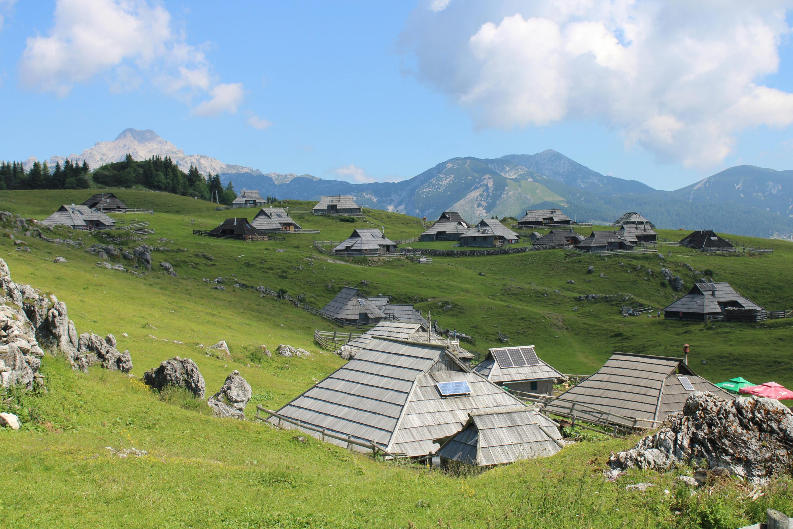 Scenic view of traditional wooden huts in Velika Planina against a backdrop of mountains and blue sky.